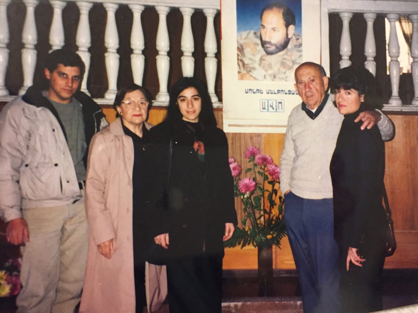 Five people in a dormitory lobby standing in front of a portrait of Monte Melkonian in fatigues